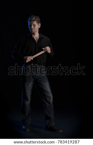 A young man in black clothes playing with drumsticks and posing on a black background with blue light