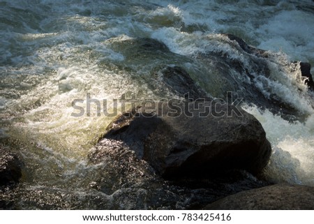 Close up on flowing river water, with sunlight highlighting rough surface riffles and foam, and waves bubbling on a dark wet boulder