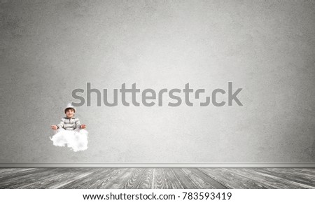 Young little boy keeping eyes closed and looking concentrated while meditating on cloud in the air with gray concrete wall on background.