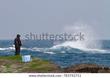 A rock fishing lover is waiting the fish and standing on the dangerous forefront of the tidal flat with green seaweed adhered on the coast. The strong wind also makes the beautiful white spray.