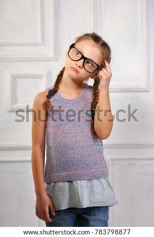 Happy thinking kid girl in fashion glasses and blouse with serious emotional face scratching the head on blue studio background