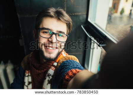 Handsome smiling hipster man blogger in glasses with beard taking selfie near window in modern loft dark interior.