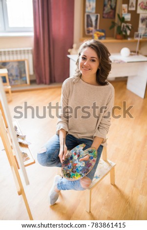 smiling young caucasian woman artist sitting with pallet in hands and canvas in art studio