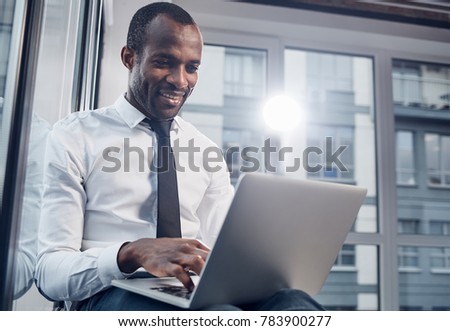 Daily life. Low angle of delightful stylish young african businessman is smiling while sitting on windowsill and typing text on laptop. Window in background with copy space in the right side