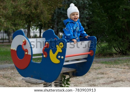 Little child in a blue jumpsuit commands standing on a ship-shaped spring rocker. Boy playing at playground outdoor