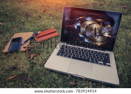 A horizontal image of a laptop viewing 3D Illustration of Bitcoin logo on screen, with a tablet, notepad, pen and mobile phone lying on grass.