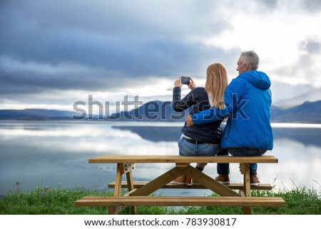 Couple sits on the bench in front of lake with mountains on the background, beautiful nature in Iceland. Woman takes a picture on the phone