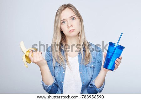 Studio shot of furious young female tired from diets has to eat banana and drink water instead of junk food. Blond European lady dreaming about pizza and soda having her healthy lunch on the go. 