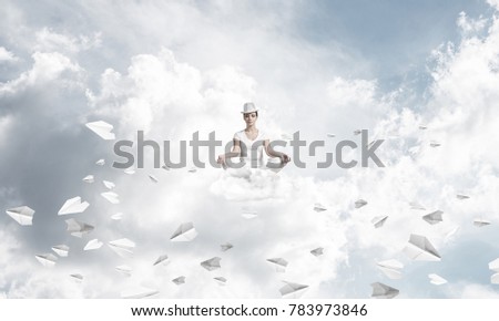 Woman in white clothing keeping eyes closed and looking concentrated while meditating among flying paper planes in the air with cloudy skyscape on background.