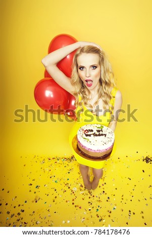 One beautiful emotional young happy blonde woman with long curly hair holding birthday cake with candles near bunch of red balloons with confetti indoor in studio on yellow backdrop, vertical picture