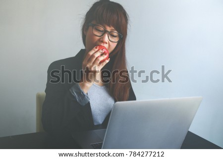 Happy asian woman eating an apple and using a laptop in an office 