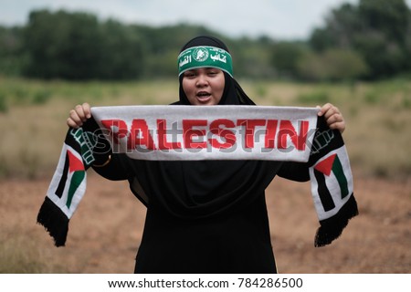 A young Muslim female teenager wearing hijab and headscarf with Arabic sign meaning "Brigade AlQassam" which related to an army group under Hamas, while holding a Palestine sign mafla