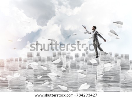Man in casual wear keeping hand with book up while standing among flying paper planes with cloudly skyscape on background. Mixed media.