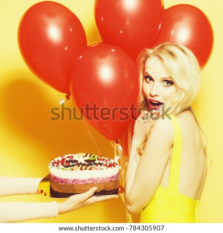 One attractive smiling young happy blond woman with long curly hair with birthday cake with candle in female hand near bunch of red party balloons in studio on yellow backdrop, square picture