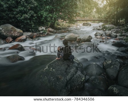 Travel image of a man sitting on the rock while looking at nice scenery of a river with motion blur slow shutter effect. The river water is blurry cause of the effect.