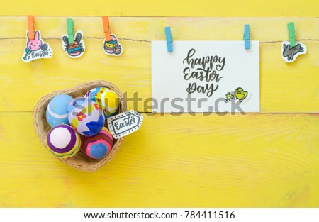 Happy Easter Day card concept. Basket of hand painted Easter egg for Christ resurrection, calligraphy handwriting card, doodles of rabbit for fertility and colorful eggs hanging on rope on wood table.