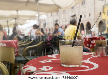A glass of cold lemon non-alcoholic drink with cubes of ice on a red tablecloth in  cafe on a historic street. 
