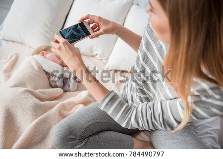 Top view of woman holding cellphone. She is taking picture of little sleeping daughter. Focus on hands with gadget