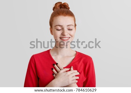 Positive redhead smiling female closes eyes and has cheerful expression, keeps hands on chest or heart, expresses positiveness and her sympathy, isolated over white background. My heart is for you