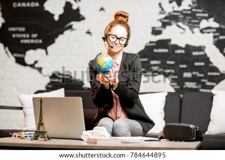 Portrait of a female travel agent in the suit and headset sitting indoors on the world map background