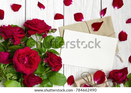blank greeting card and envelope with red roses flowers, petals and gift box over white wooden table. valentines day background. top view. mock up