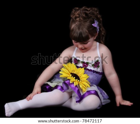 sweet little girl in purple and white ballet outfit sitting looking down at sunflower. isolated on black