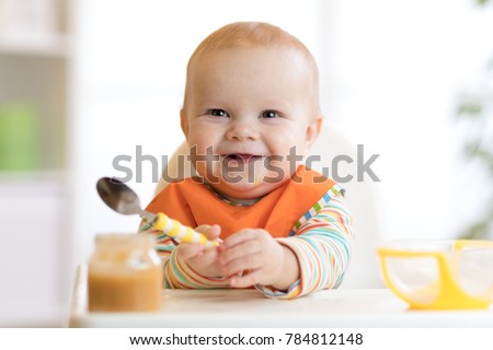 Cheerful baby child eats food itself with spoon. Portrait of happy kid boy in high chair. Royalty-Free Stock Photo #784812148