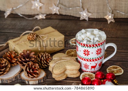 on a brown wooden background lie a Christmas present, pine cones, ginger biscuits, a cup of cocoa with marshmallow, dried lemon slices and Christmas toys
