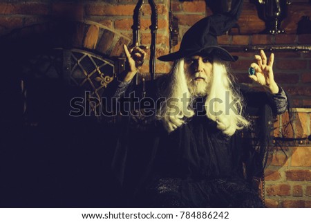Old man wizard with long gray hair beard in black costume and hat for Halloween holding blue gem stone and silver pendant for hypnosis on wooden background