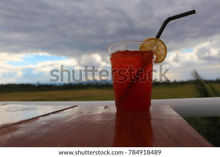 A glass of cool lemon tea on a wooden table. There are trees and fields as the background.