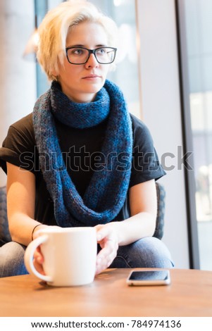 Young beautiful blonde woman in black glasses and in a knitted dark blue scarf sitting near window in modern city coffee shop