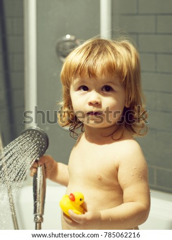 Cute happy smiling funny undressed boy child with blonde curly wet hair taking shower in bath with water indoor playing with toy yellow color duckling, vertical picture