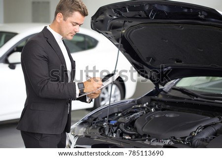 Shot of a professional car salesman working at the dealership standing near a new auto with an open hood signing documents copyspace sales consumerism assistance manager occupation career job.