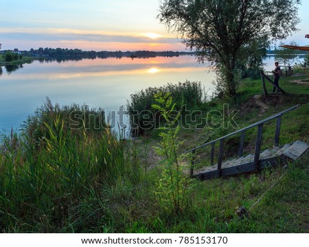 Happy woman tourist with compact photo camera on picturesque summer sunset lake calm beach with camping pitch on shore. Concept of tranquil country life, eco friendly tourism, camping, fishing.
