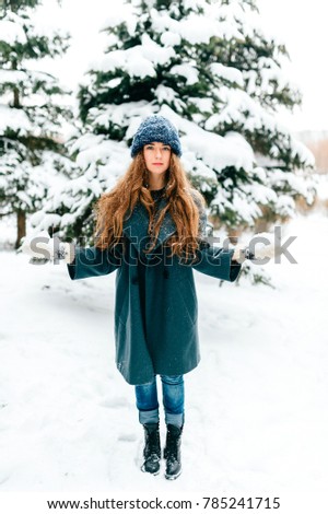 Young beautiful teenager girl in oversized wool coat with long brown beautiful hair standing in winter city park with snowy spruces on background. Fashionable stylish lady with amazaing blue eyes