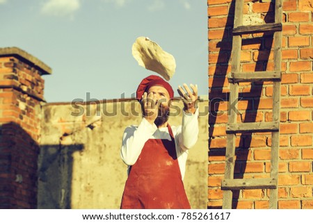 Man cook chef hipster with long beard on handsome face in red hat and white uniform throwing dough up on blue sky ladder and brick wall background outdoor