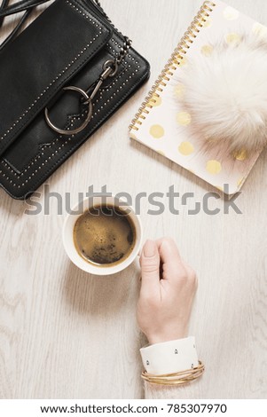 Top view woman hand holding cup of black coffee on flat lay. Workspace with notebook in pink and gold, fashion handbag and fur ball