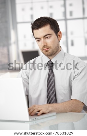 Young man working on laptop in bright office sitting at desk.?