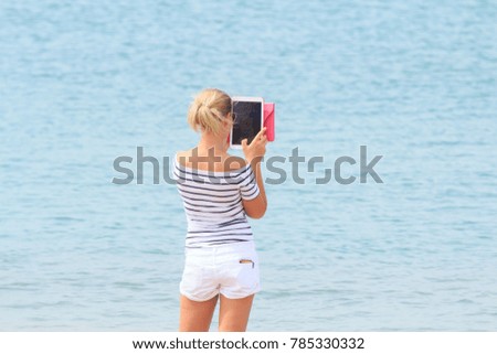 Lifestyle of Blond Hair Woman Tourist is Holding Computer Tablet for Take Photo on Sea Beach. Copy Space for Text.