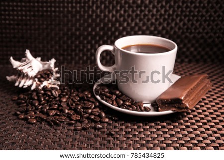 A cup of coffee with a chocolate bar, scattered coffee beans and a shell.