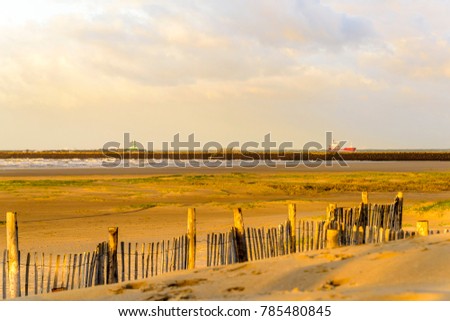 Early morning at stormy sea with blue skies, puffy clouds, sand dunes, beachgoers with wooden fence and ship  