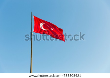 Turkish flag on long metal iron pole waving in blue sky. Red flag featuring a white star and crescent. Flag is often called al bayrak  and is referred to as al sancak in the Turkish national anthem.