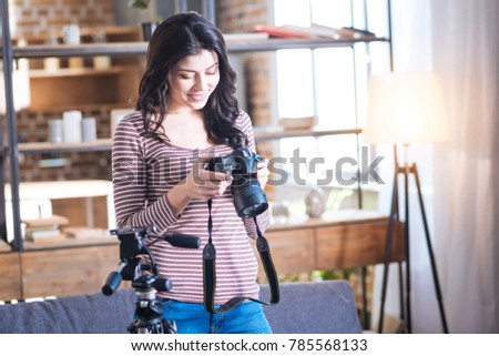 Modern camera. Delighted nice young woman standing in the room and holding a camera while looking at the photos