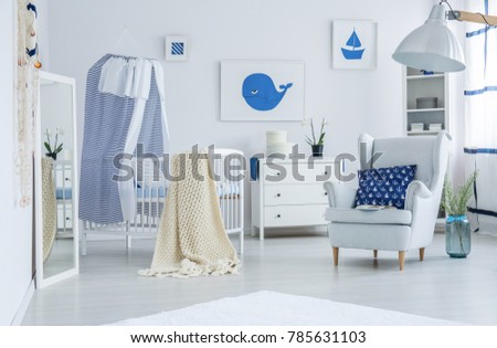 Blue pillow with nautical pattern lying on a white armchair next to wooden crib with beige blanket prepared for a newborn baby