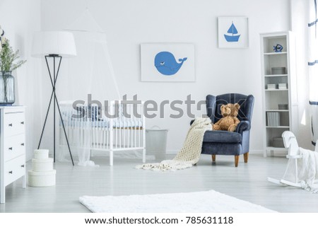 White crib standing between a navy blue armchair and oversized lamp in cozy nursery with nautical design