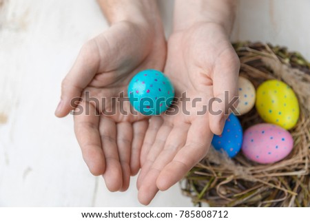 Close-up of man's hands is holding cyan egg above the nest with colorful Easter eggs on a rustic background. Easter concept.