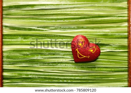 The heart is red with a smile on the background of woven green leaves with drops of dew