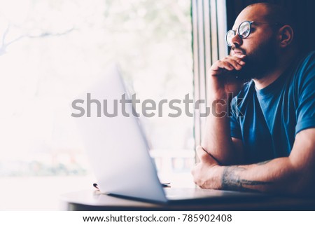 Handsome contemplative male entrepreneur dressed in casual clothing thinking about creative ideas for upcoming startup project.Young hipster guy in spectacles pondering during sitting indoors