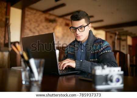 Young attractive hipster man in glasses and winter sweater working on laptop in cafe. Freelancer businessman browsing internet at coffeshop. Photographer retouching photos. Vintage camera on table.