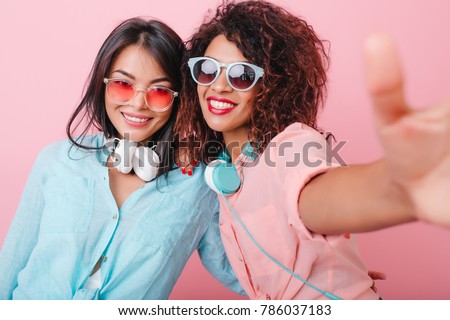 Two tanned ladies with headphones making selfie during indoor party. Enthusiastic curly woman in sunglasses taking picture of herself near romantic asian girl in blur cotton shirt.